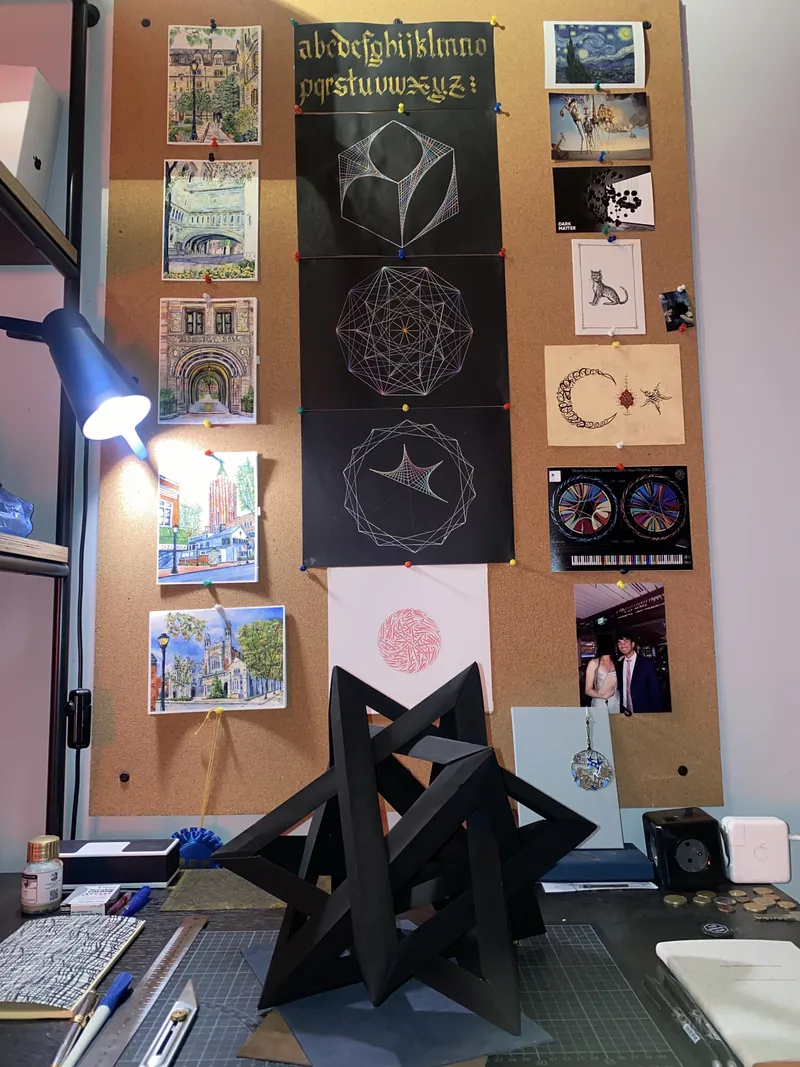 Drafting desk below a corkboard of calligraphy, geometric sketches and postcards, with a folded paper sculpture on the cutting mat